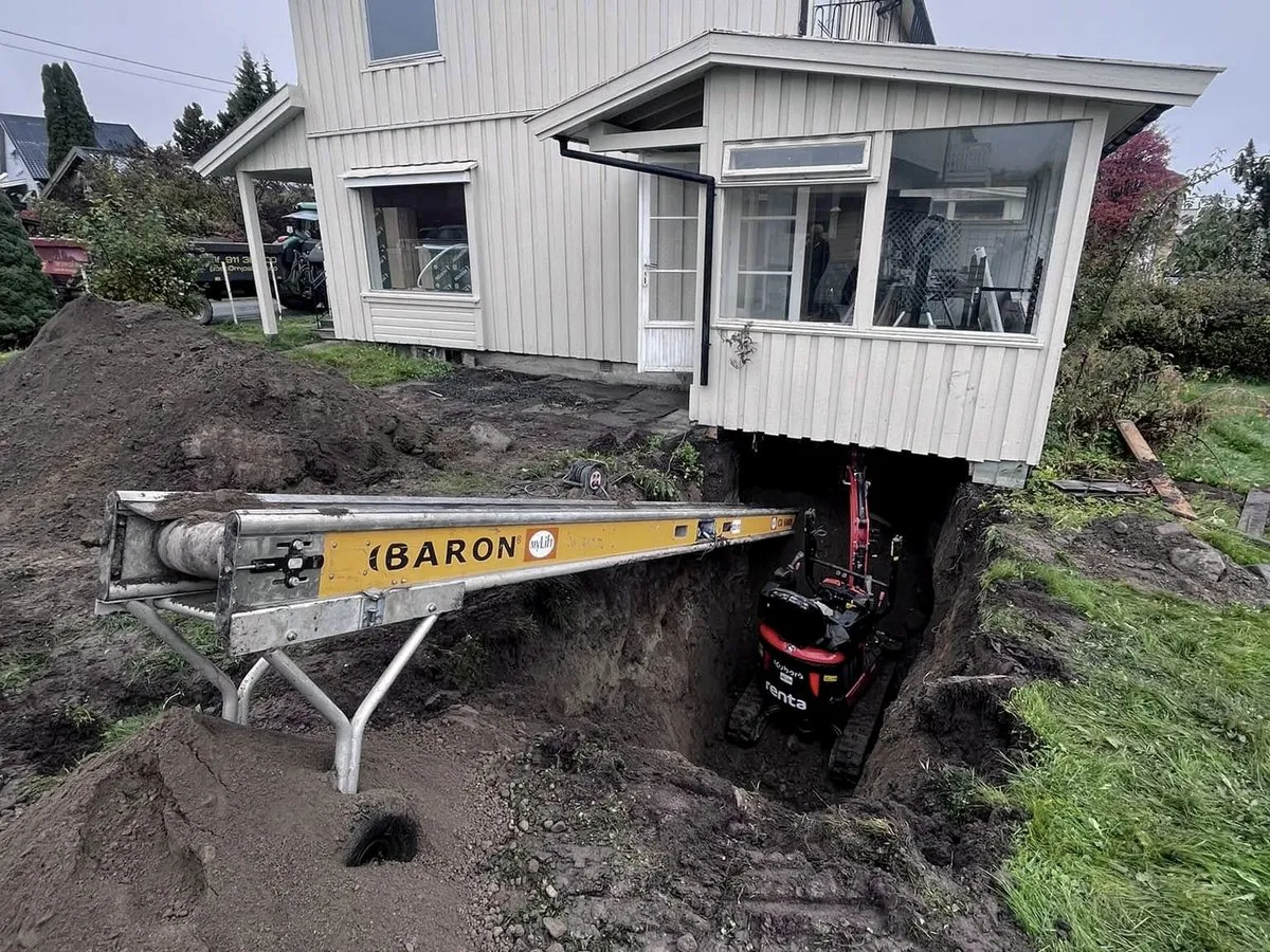 Transportbånd og minigraver under et hus der det graves for ny drenering