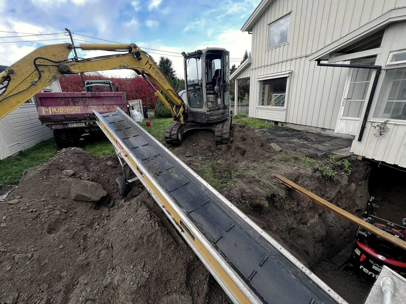 Gravemaskin og transportbånd ved et hus der det graves for ny drenering